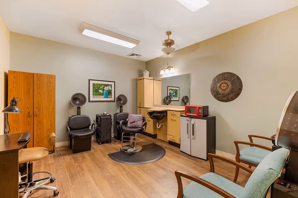 Interior view of a hair salon room with wooden flooring, two black salon chairs with hair dryers, a salon styling chair in front of a mirror, a wooden cabinet, a small refrigerator with a red microwave on top, and two green cushioned chairs along the wall.