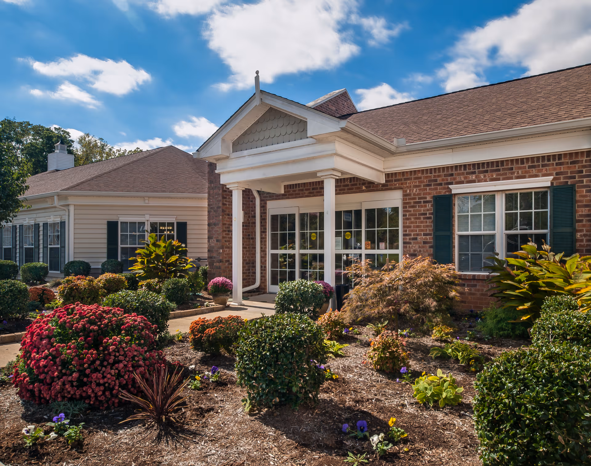 Exterior view of Charter Senior Living of Paris showing a brick building entrance with white columns and a small covered porch. The foreground features a landscaped garden with various shrubs, flowering plants, and mulch under a partly cloudy blue sky.
