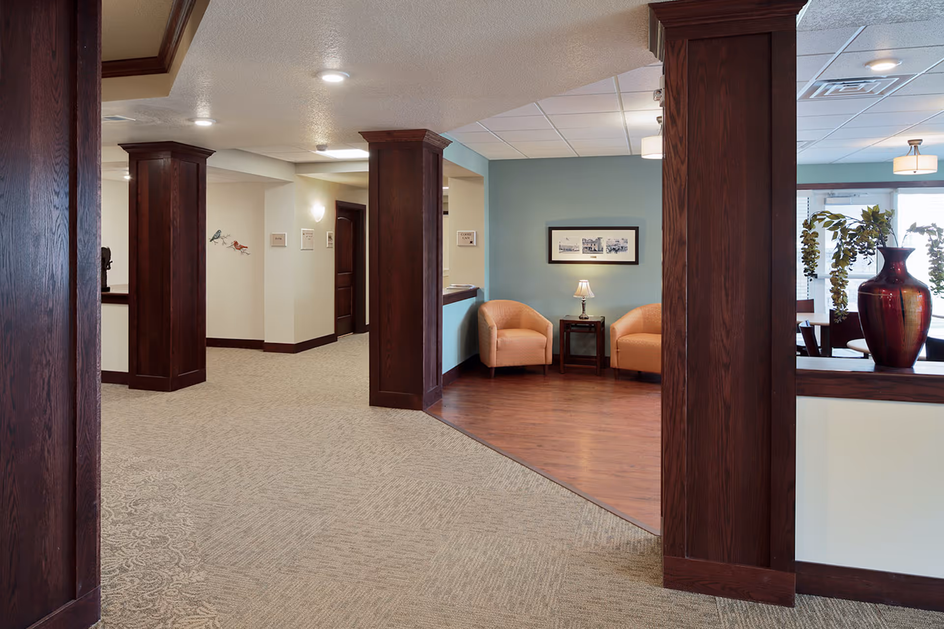 Interior view of a senior living facility hallway with dark wooden columns and trim. To the right, there is a small seating area with two beige armchairs, a side table with a lamp, and a framed picture on a light blue wall. The floor transitions from carpet in the hallway to wood in the seating area. A large red vase with greenery is visible on a ledge near the seating area.