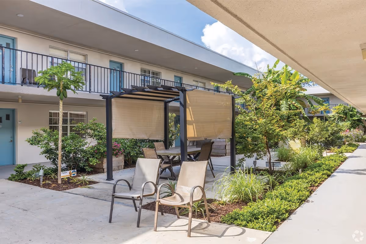 Outdoor courtyard area of a senior living community featuring a shaded pergola with a table and chairs underneath, two additional chairs in the foreground, surrounded by various green plants and small trees, with a two-story building with blue doors and windows in the background under a partly cloudy sky.