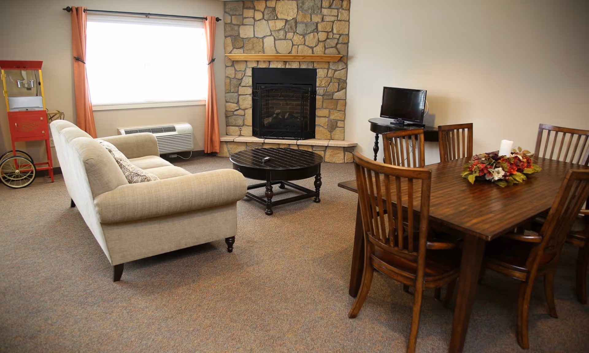 A cozy living room with a beige sofa facing a stone fireplace. There is a round black coffee table in front of the sofa and a wooden dining table with six chairs to the right. A small TV sits on a side table near the dining area. A red popcorn machine is positioned near a window with orange curtains.