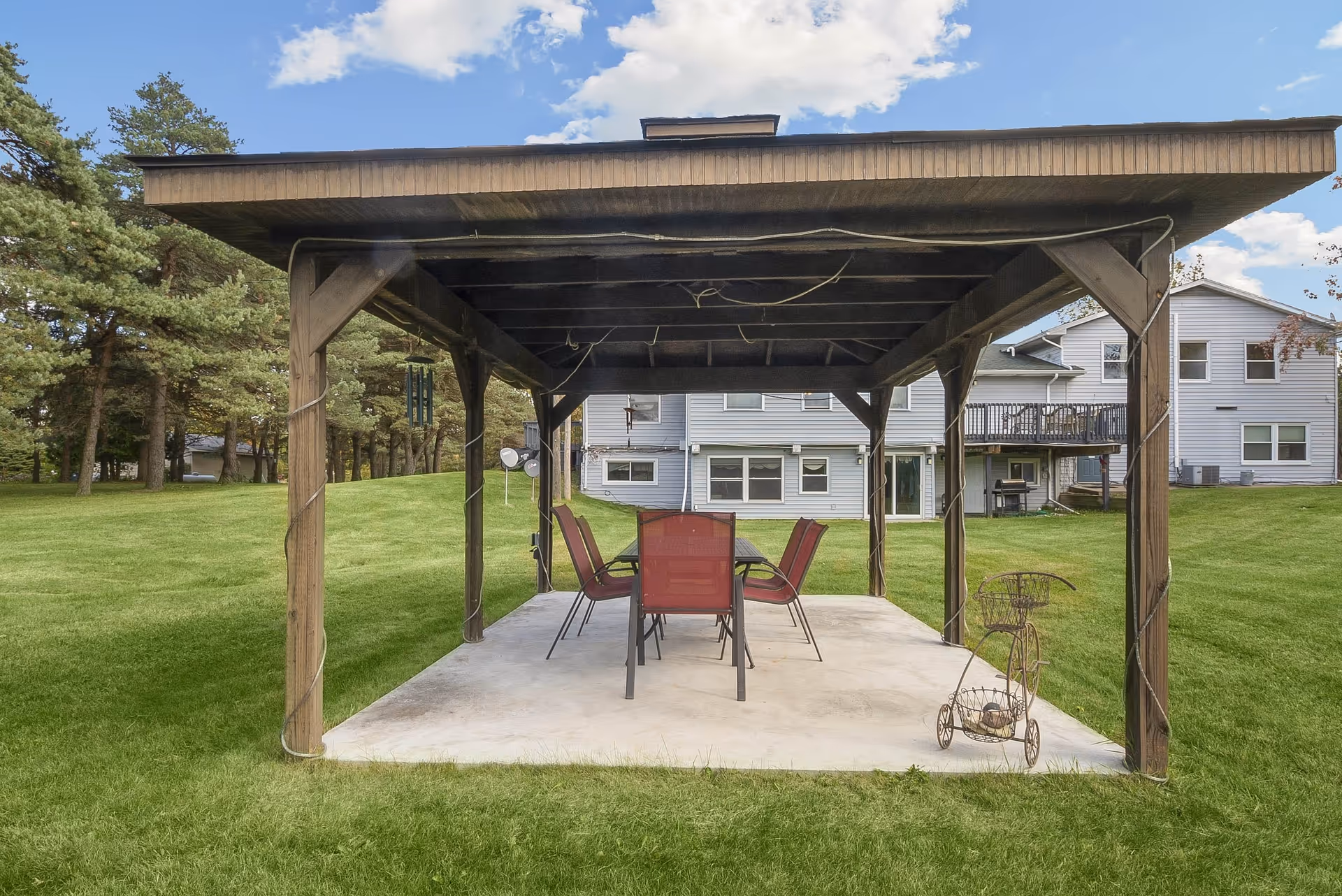 Outdoor covered patio area with a wooden pergola structure, a table, and six red chairs on a concrete slab. The patio is surrounded by green grass and trees, with a large light gray building in the background under a partly cloudy sky.