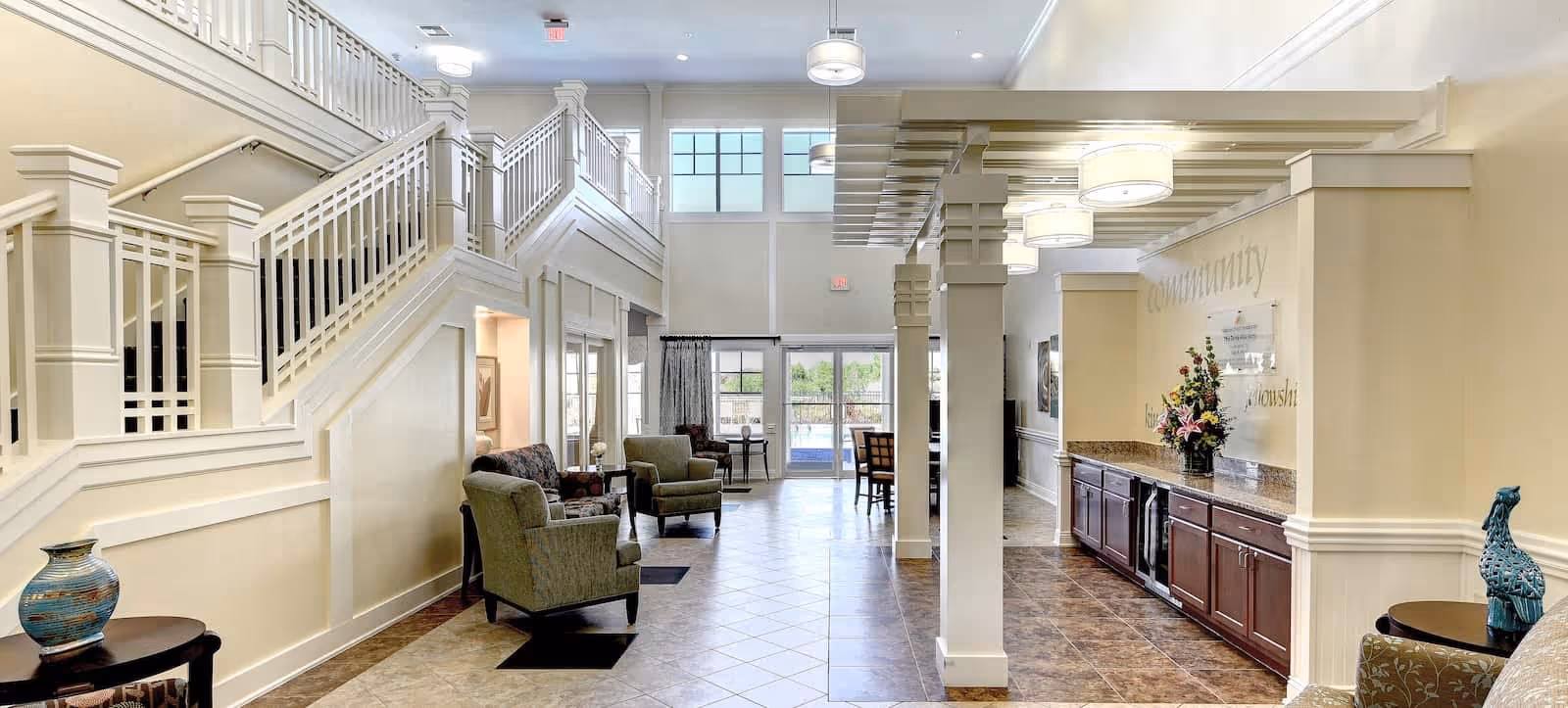 Interior view of a spacious senior living facility common area with a staircase on the left, several armchairs arranged along the wall, and a long counter with cabinets and a flower arrangement on the right. Large windows and glass doors at the far end allow natural light to fill the space.