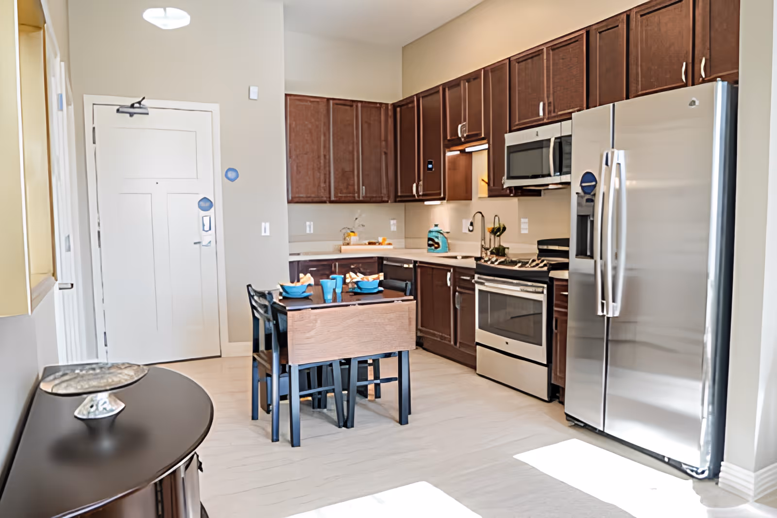 Bright modern kitchen with dark wood cabinets, stainless steel refrigerator and oven, and a small dining table set in the center.