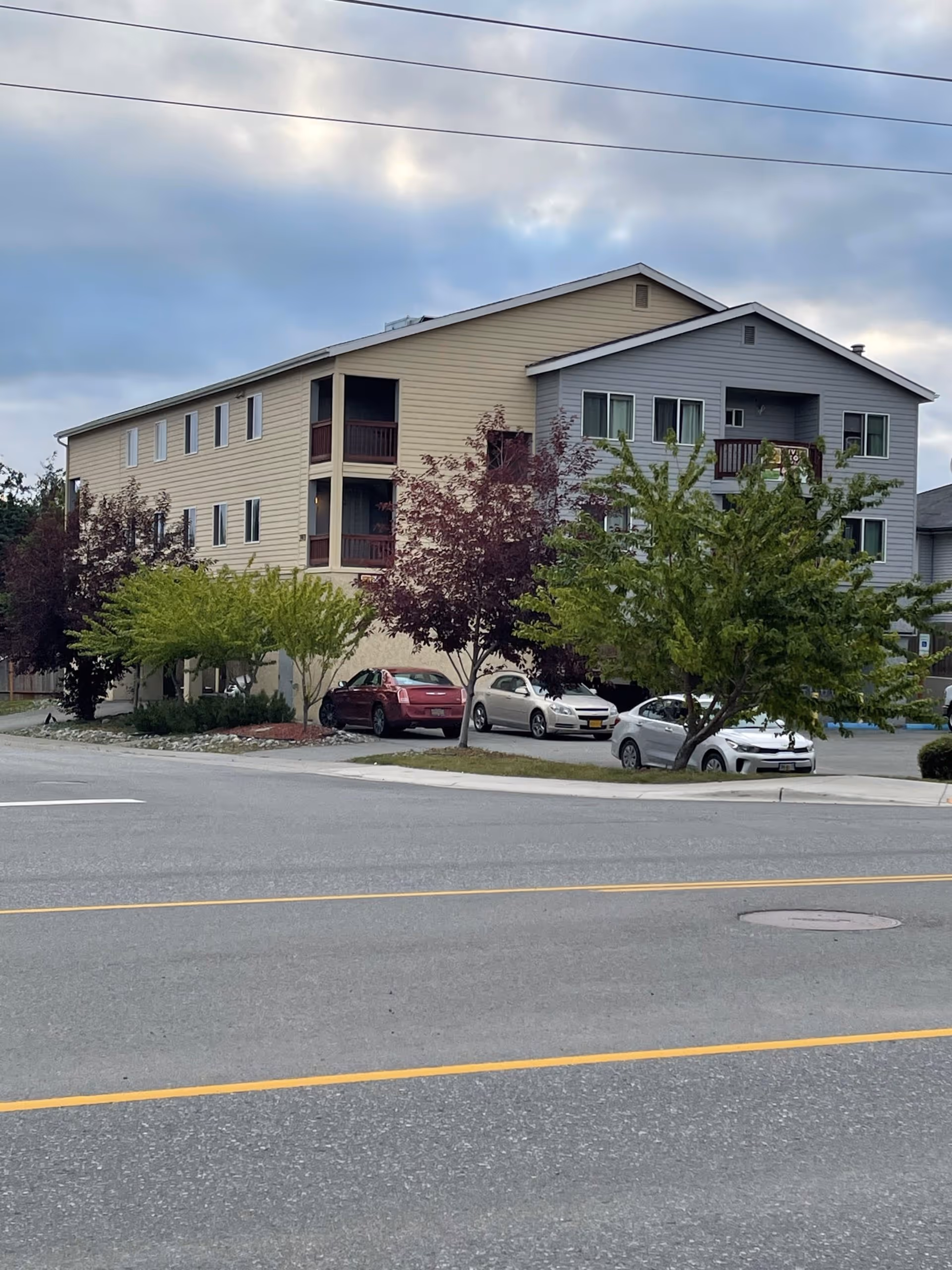 Exterior view of a multi-story residential building with beige and gray siding, surrounded by trees and parked cars under a cloudy sky.