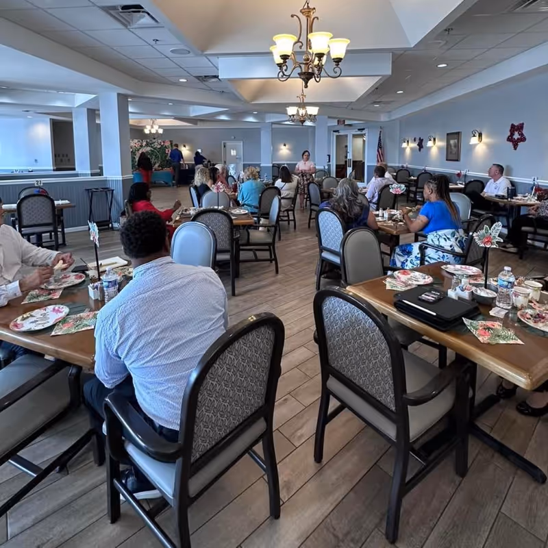 Residents and staff seated at tables in a communal dining room with chandeliers and place settings.