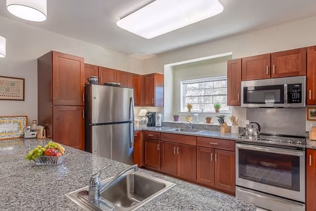 A modern kitchen with granite countertops, wooden cabinets, a stainless steel refrigerator, oven, microwave, and a sink in the foreground. There are small potted plants on the windowsill above the sink and a bowl of fruit on the counter.