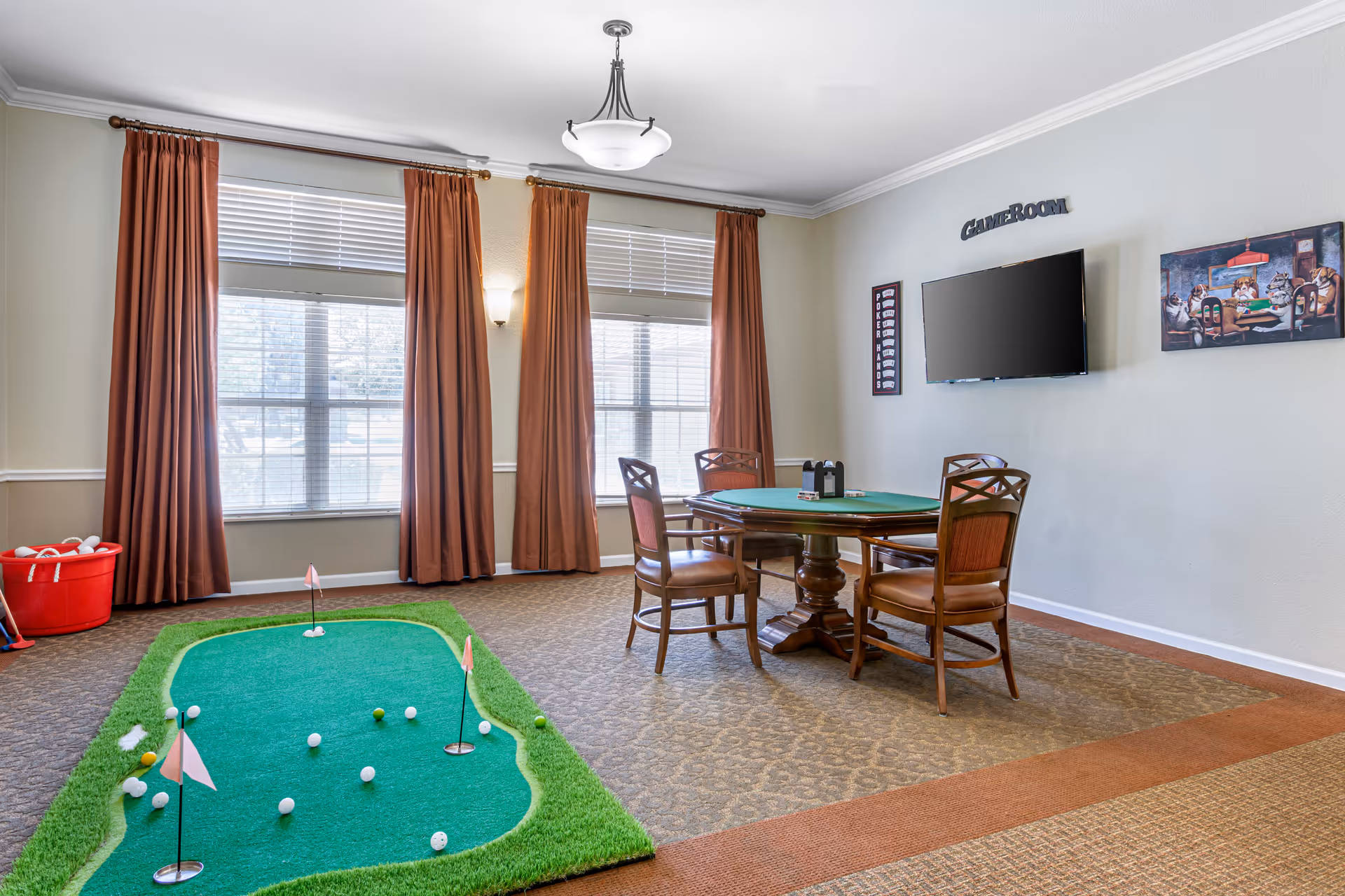 A game room with a small indoor putting green on the left side and a round card table with four chairs on the right side. The wall has a mounted flat-screen TV with the word 'GAMEROOM' above it and two framed pictures on either side. The room has large windows with brown curtains and beige walls.