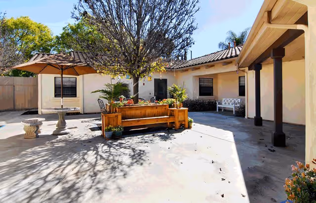 Outdoor courtyard area with a central tree surrounded by a wooden planter box with flowers. There is a round stone table with an umbrella and stone benches on the left side, and a white bench against the building wall on the right. The courtyard is paved and enclosed by a beige building with a tiled roof and a wooden fence in the background.