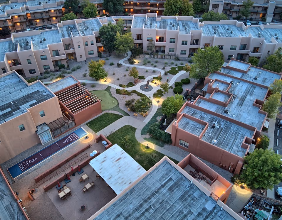 Aerial view of a courtyard with winding pathways, landscaping, patios and a shuffleboard court surrounded by stucco apartment buildings.