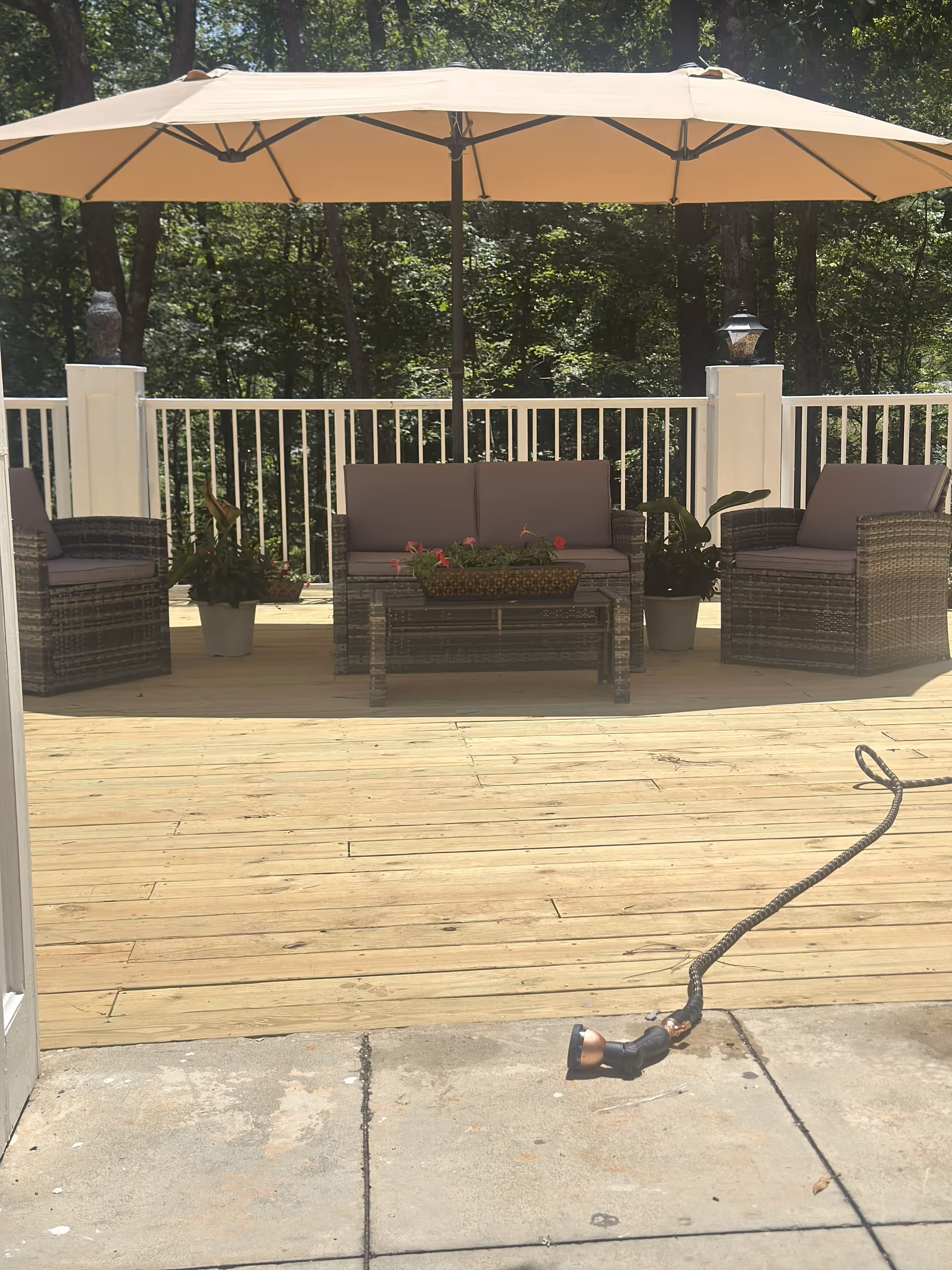 Outdoor patio area with wooden deck flooring, featuring a large beige umbrella providing shade over a wicker loveseat and two matching armchairs with cushions. There are potted plants on either side of the loveseat and a rectangular planter with flowers on the coffee table. A garden hose with a spray nozzle is lying on the concrete floor in the foreground. Trees and greenery are visible in the background beyond a white railing fence.