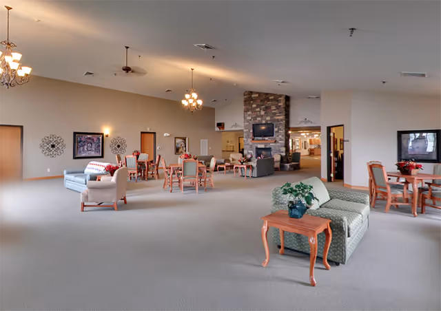 Spacious assisted-living common room with sofas, tables, chairs, and a stone fireplace under chandeliers.