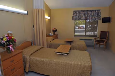 Two twin beds with beige bedding and matching wood furniture in a sunlit senior living bedroom.
