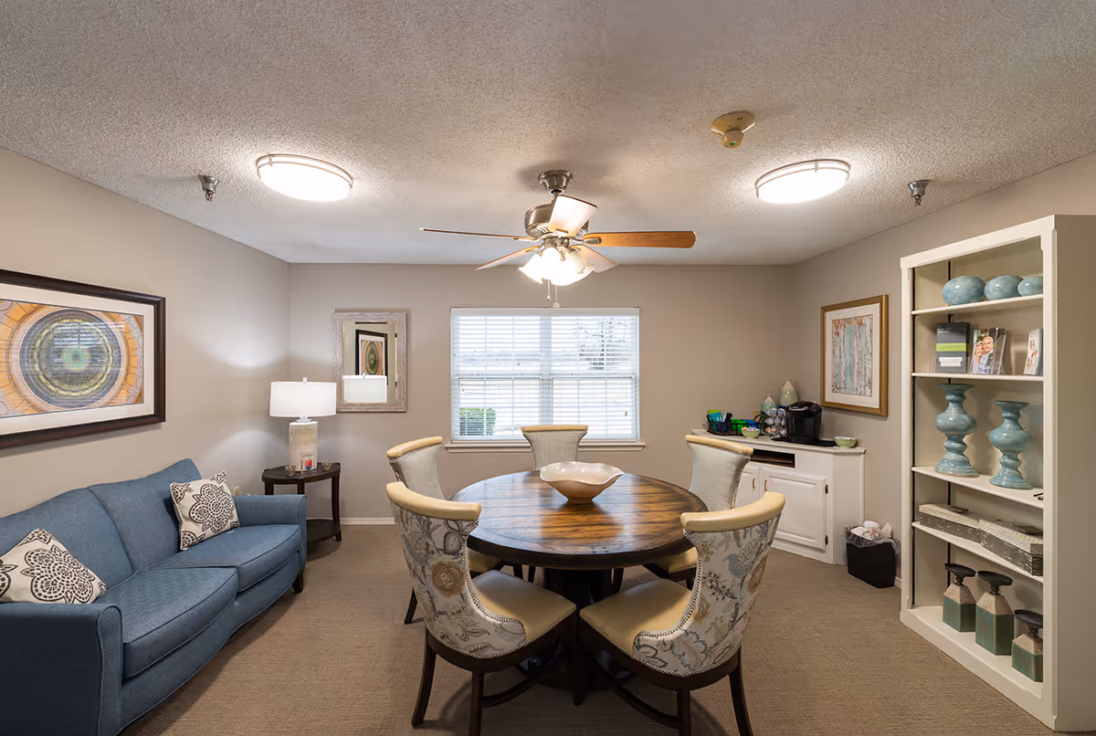 A cozy interior room with a round wooden table surrounded by six upholstered chairs with floral patterns. There is a blue sofa with patterned pillows on the left side, a small side table with a lamp, a window with blinds in the center, and a white shelving unit with decorative items on the right. The ceiling has a fan with lights and two round ceiling lights.