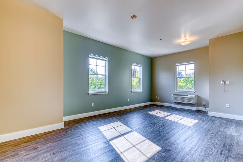 Empty room in a senior living facility with three windows letting in natural light, light green and beige walls, wood-style flooring, and a wall-mounted air conditioning unit.