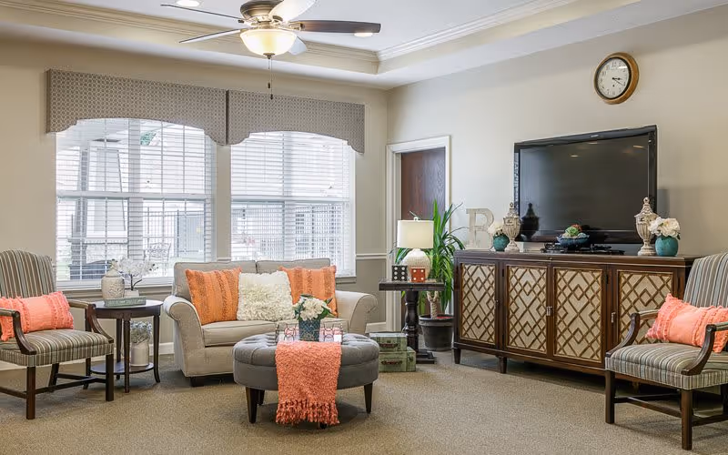 A cozy living room with a beige sofa adorned with orange and white pillows, two striped armchairs with orange cushions, a round gray ottoman with an orange throw, and a wooden cabinet with a flat-screen TV mounted above it. The room has large windows with blinds and a ceiling fan with a light fixture.
