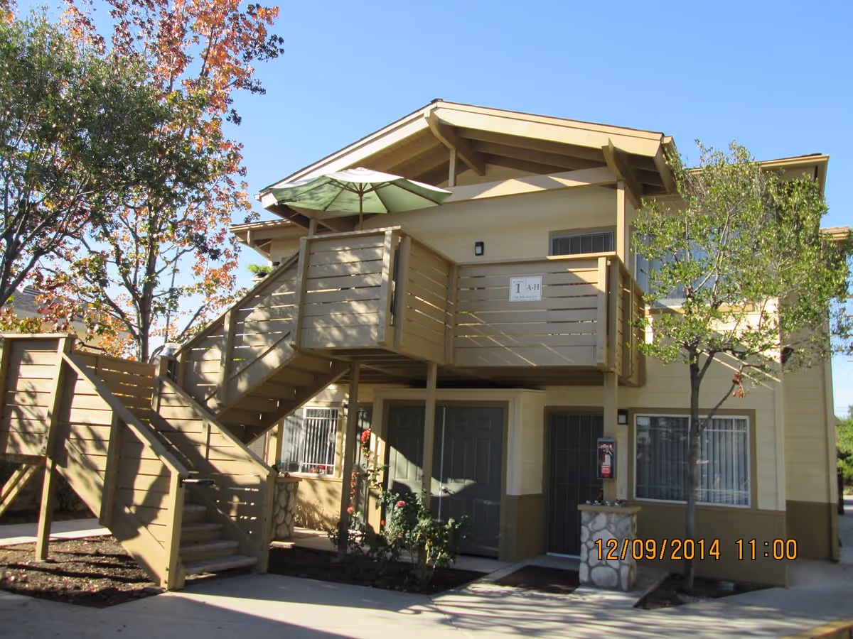 Exterior view of a two-story residential building with beige siding and brown trim. There is an outdoor staircase leading to an upper balcony with a green patio umbrella. Trees with autumn-colored leaves and green foliage surround the building. The building has multiple windows with security bars and two doors on the ground level. The date and time stamp on the image reads 12/09/2014 11:00.