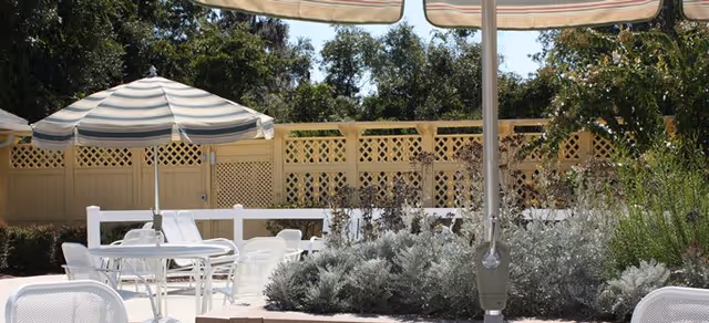 Outdoor seating area with white metal tables and chairs, striped umbrellas providing shade, surrounded by greenery and a lattice fence in the background.