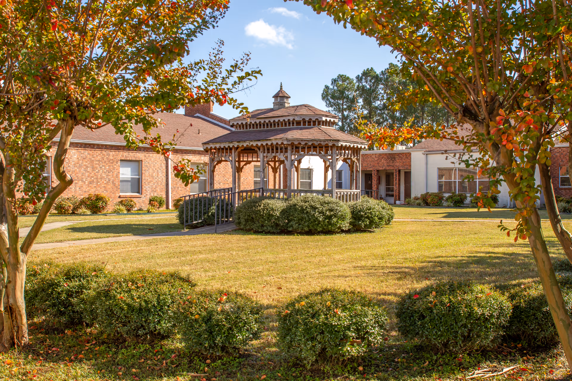 Outdoor view of a senior living facility courtyard featuring a wooden gazebo surrounded by bushes and trees with autumn foliage. The building is made of brick and has multiple windows, with a clear blue sky overhead.
