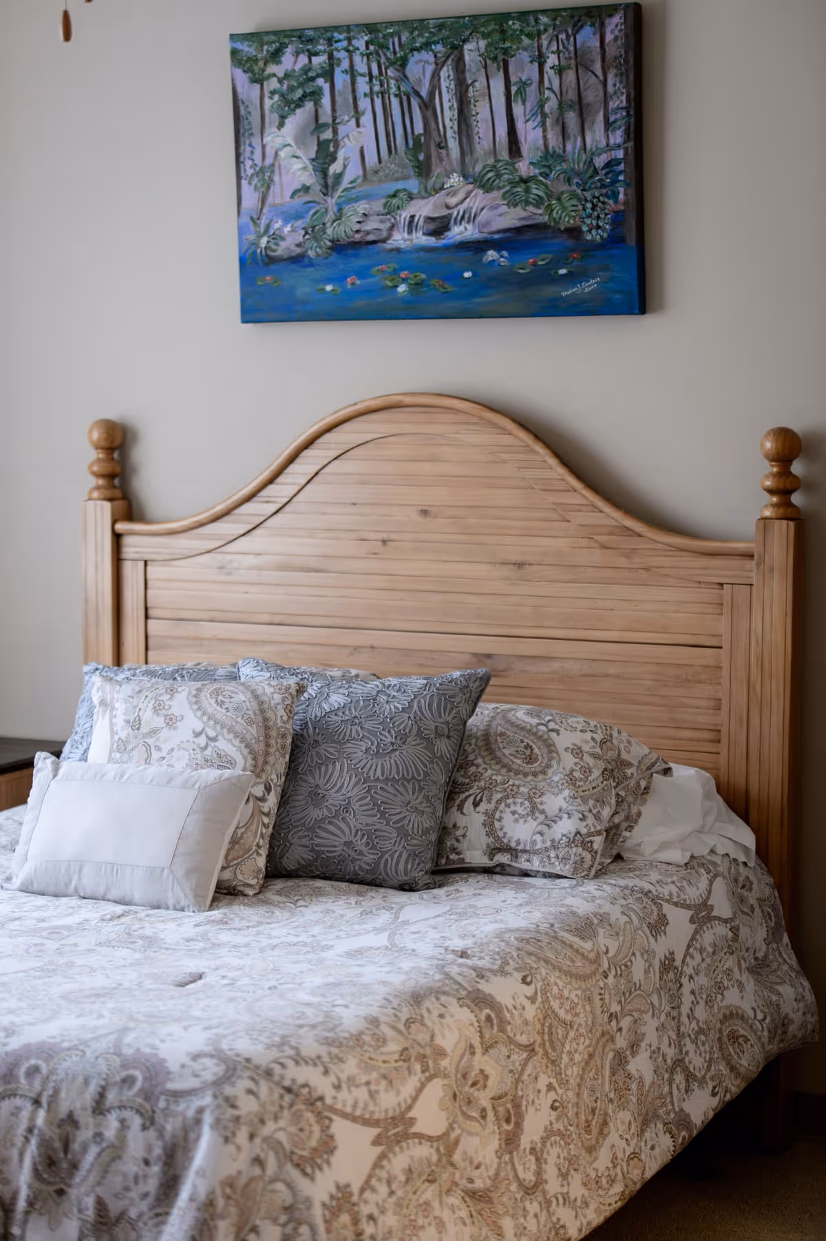 A wooden bed with a curved headboard, adorned with multiple pillows in various patterns and colors, including gray and paisley designs. Above the bed hangs a painting depicting a serene forest scene with a small waterfall and lily pads on a pond.
