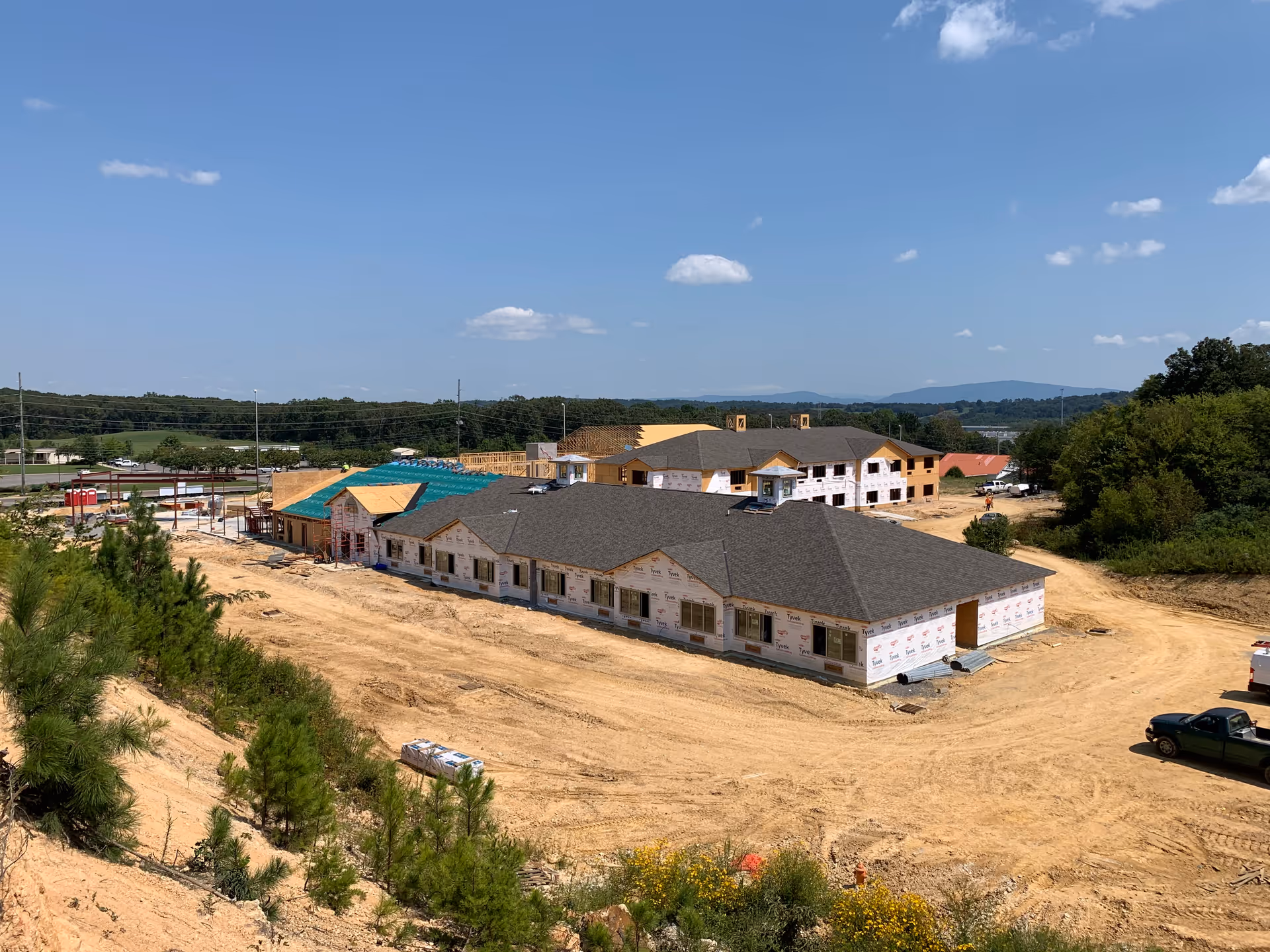 Aerial view of multiple senior living buildings under construction on a dirt lot with surrounding trees and distant hills.