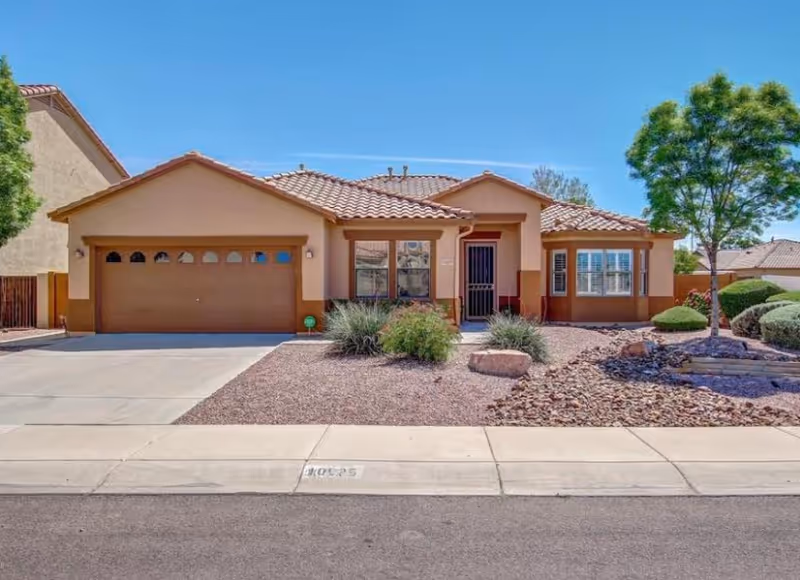 Single-story house with a tiled roof, a two-car garage, desert landscaping with rocks and bushes, and a tree on the right side under a clear blue sky.