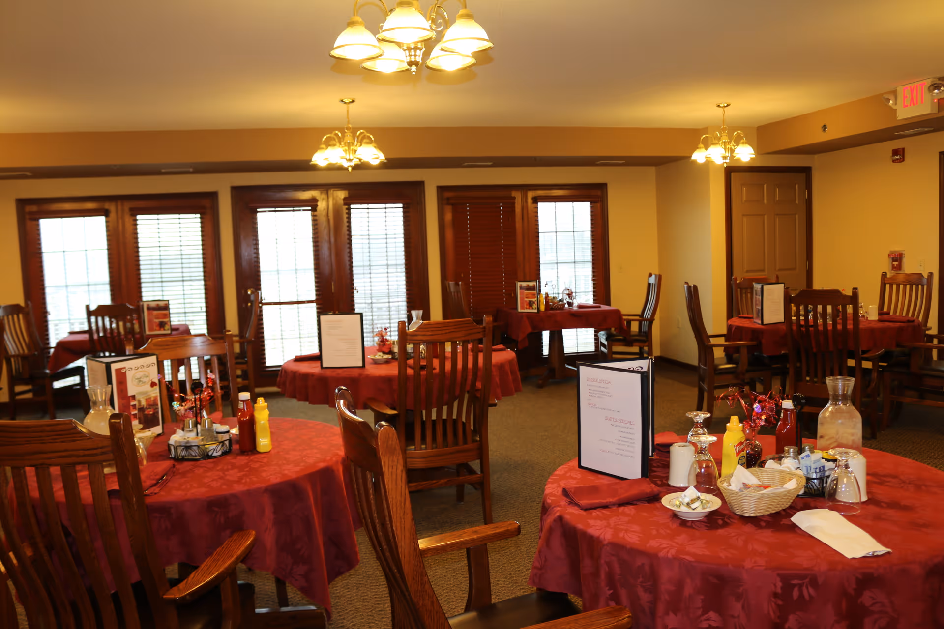 Dining room with round tables covered in red tablecloths, wooden chairs, menus and condiments on the tables.