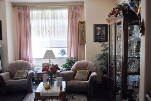 A cozy living room with two cushioned armchairs facing a wooden coffee table adorned with a vase of red flowers and decorative items. Behind the chairs is a large window with sheer white curtains and pink drapes. To the right, there is a tall glass-fronted wooden cabinet displaying glassware and decorative items. The room also features a table lamp, framed artwork on the wall, and several potted plants.