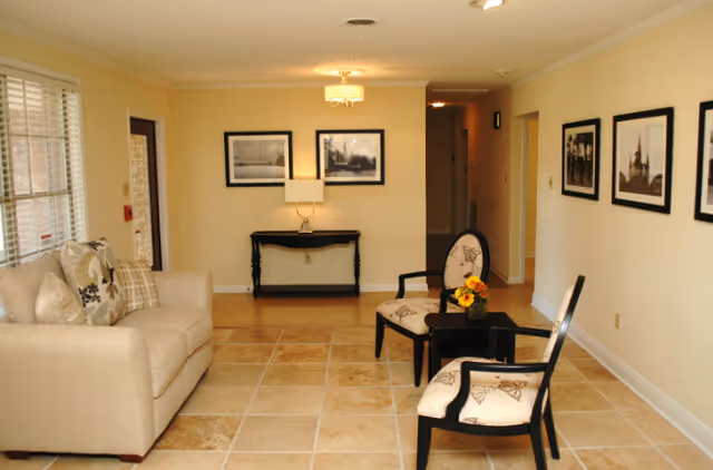 A cozy living room area with a beige sofa adorned with patterned cushions on the left, two upholstered chairs with butterfly designs around a small black table with a flower arrangement in the center, beige tiled floor, cream-colored walls decorated with framed black and white photographs, a black console table with a lamp against the far wall, and a hallway leading further into the facility.