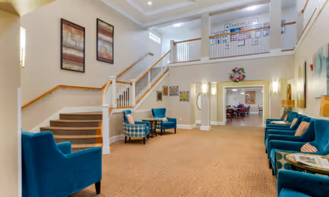 Interior view of a senior living facility lounge area with blue upholstered chairs arranged along the walls and near a staircase. The space has beige walls, carpeted floors, framed artwork on the walls, and a railing on the upper level. There is a doorway leading to another room with tables and chairs visible in the background.