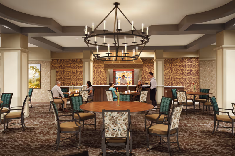 Spacious dining room with round wooden tables and patterned chairs, a large chandelier overhead, and staff serving seated residents.