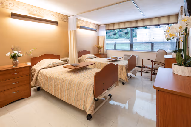 A bright and clean room with two hospital-style beds, each with beige patterned bedspreads and wooden headboards and footboards. Each bed has a small overbed table with a book on it. There is a wooden nightstand with a vase of flowers between the beds. The room has large windows with blinds, letting in natural light. A wooden dresser with a potted orchid and a chair are also visible. The walls are painted a warm beige color with decorative molding near the ceiling.
