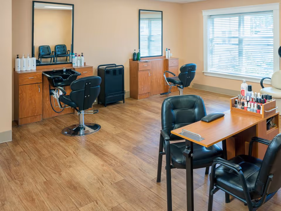 Interior view of a salon area in a senior living facility with two black salon chairs in front of mirrors and wooden cabinets, a manicure table with two black chairs and a display of nail polish bottles, and a large window with blinds letting in natural light.