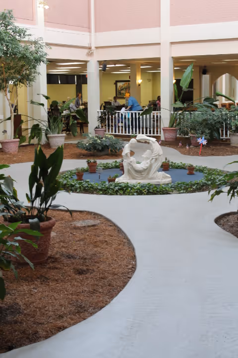 Indoor courtyard area with a circular garden bed in the center containing a white statue surrounded by small potted plants. The courtyard has a curved walkway and several large potted plants. In the background, there are people sitting and interacting in a common area with white railings and columns.