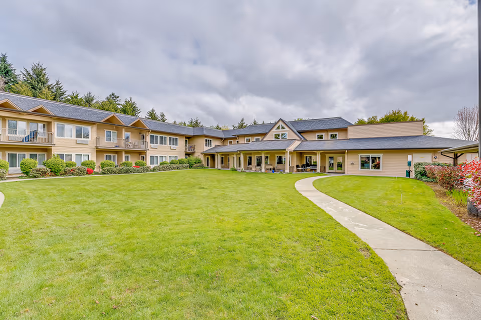 Two-story senior living building with a covered central entrance facing a large grassy courtyard and curved walkway under a cloudy sky.