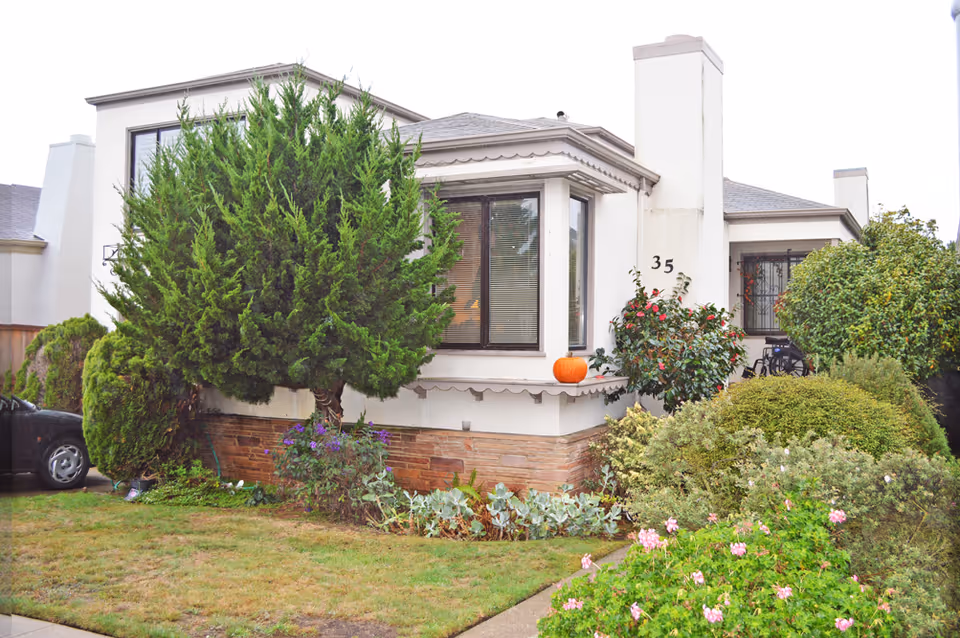 Exterior view of a single-story residential building with white walls and a stone base. The house has large windows, a chimney with the number 35, and is surrounded by various green bushes, trees, and flowering plants. A pumpkin is placed on the ledge near the window.
