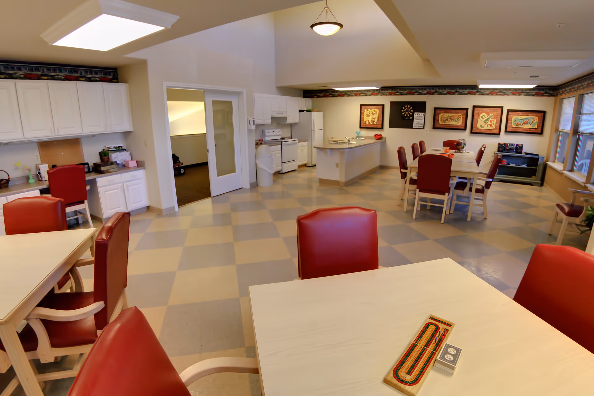 A spacious common area in a senior living facility featuring a kitchen with white cabinets, stove, and refrigerator. There are multiple tables with red cushioned chairs arranged for dining or activities. The floor has a checkered pattern, and the walls are decorated with framed vintage-style posters. The room is well-lit with ceiling lights and natural light from windows on the right side.