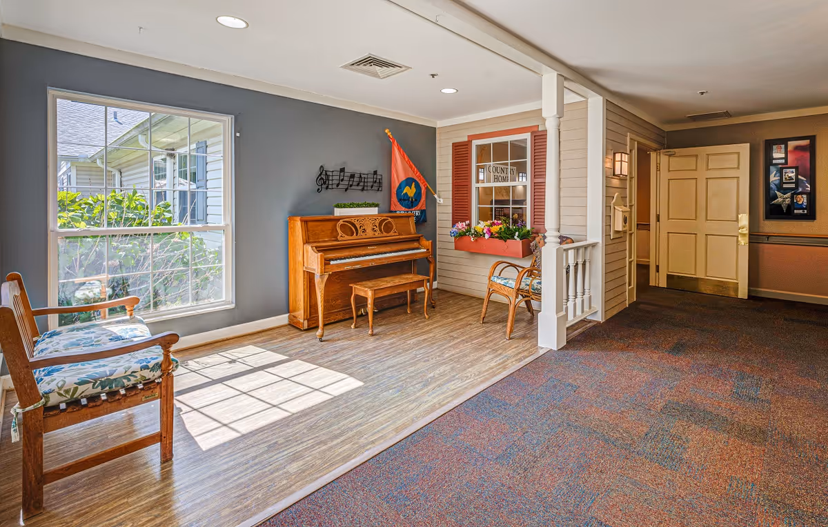 A bright and cozy interior corner of a senior living facility featuring a wooden piano with a matching bench, two cushioned wooden chairs with floral upholstery, a large window letting in natural light, a decorative flag, a wall-mounted musical note decoration, and a faux window with flower boxes labeled 'Country Home'. The floor is a combination of wood and carpet, and there is a hallway with an open door in the background.