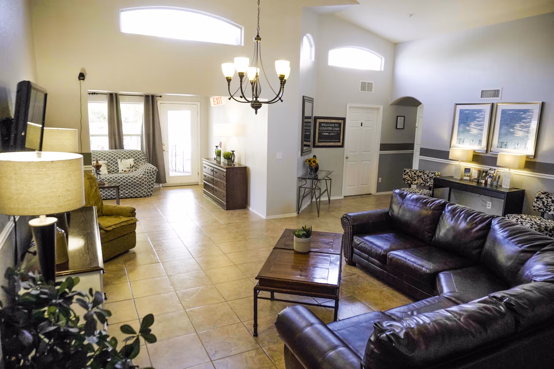 A bright and spacious living room area with a large dark leather sectional sofa, a wooden coffee table with a small plant, and two patterned chairs near a console table with lamps and framed pictures. The room has tiled floors, high ceilings with a chandelier, and large windows letting in natural light. There is a doorway leading to another room and a door with an exit sign above it.