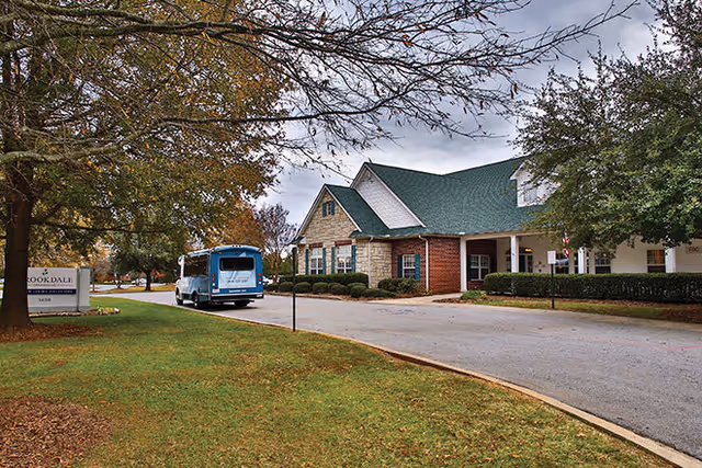 Exterior view of a senior living facility building with a green roof and brick and stone facade. A blue shuttle bus is parked near the entrance. Trees with autumn foliage surround the area, and a sign for Brookdale is visible on the left side of the image.