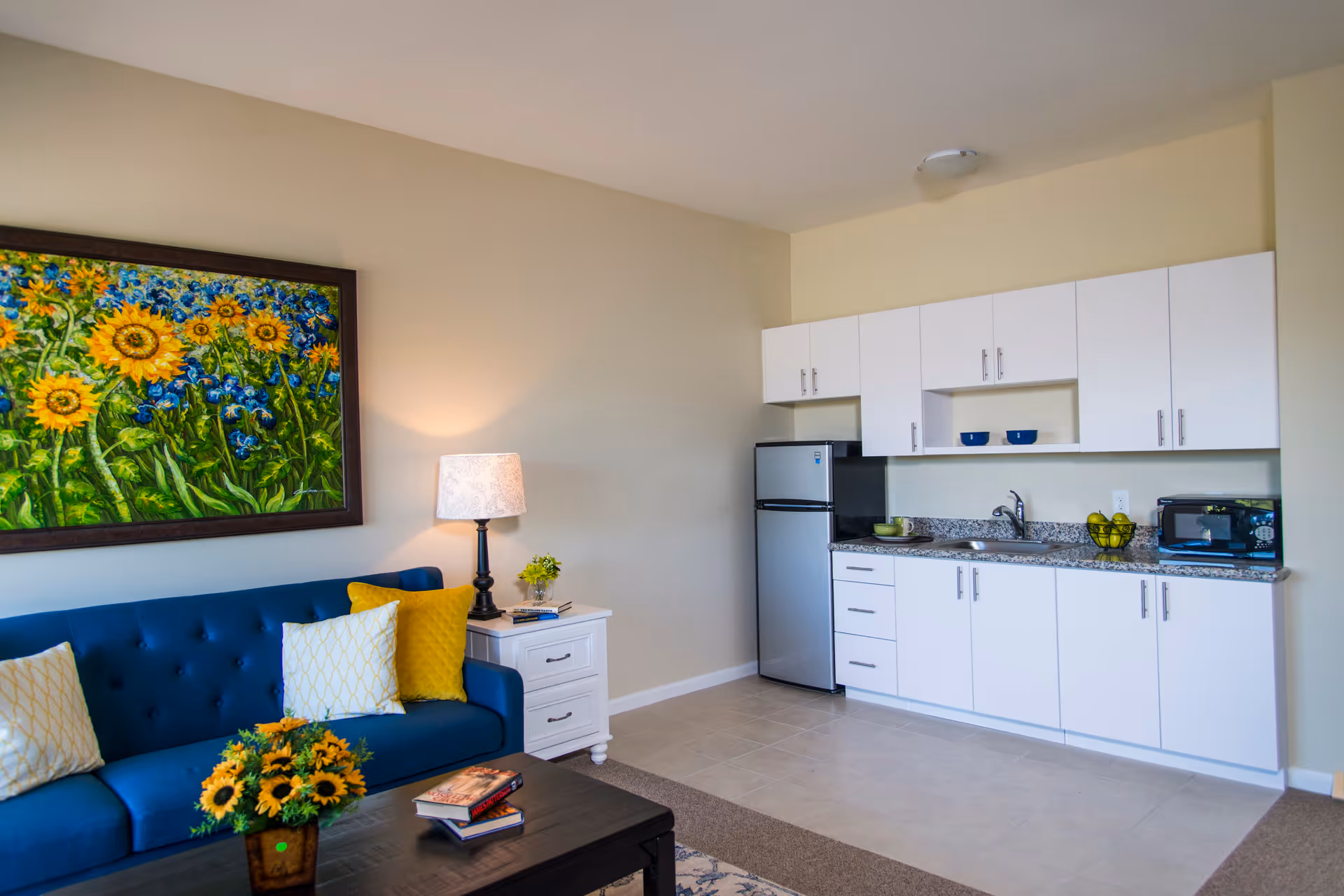 A cozy living area with a blue sofa adorned with yellow and white pillows, a white side table with a lamp and small plant, a coffee table with a sunflower bouquet and books, and a kitchenette with white cabinets, a small refrigerator, a microwave, and a granite countertop. A colorful painting of sunflowers and blue flowers hangs on the wall above the sofa.