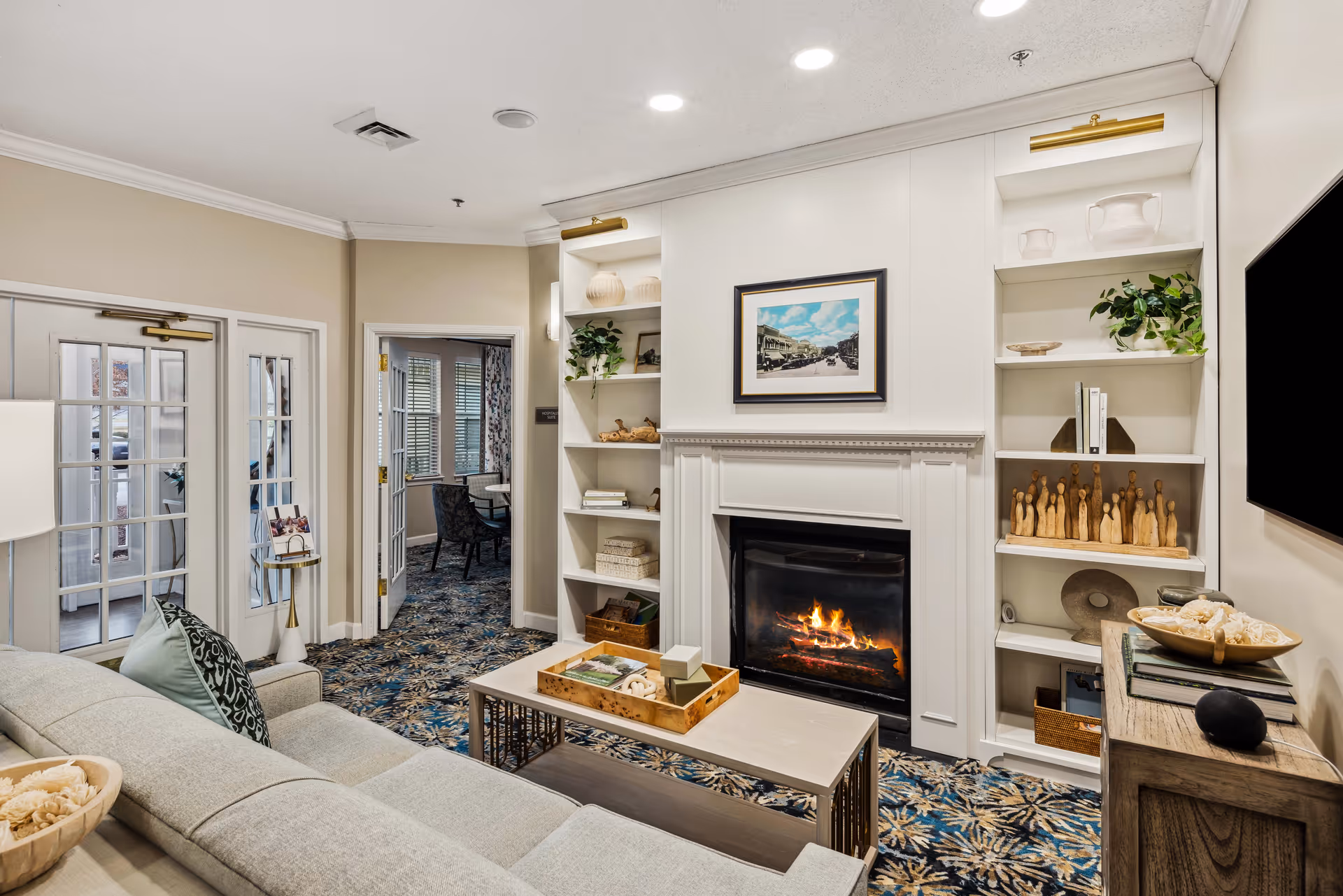 A cozy living room in TerraBella Lincolnton featuring a light gray sofa with decorative pillows, a coffee table with a wooden tray, a lit fireplace with white mantel, built-in shelves with decorative items and plants, a large flat-screen TV mounted on the wall, and a patterned carpet. French doors and an open doorway lead to another room with a table and chairs.