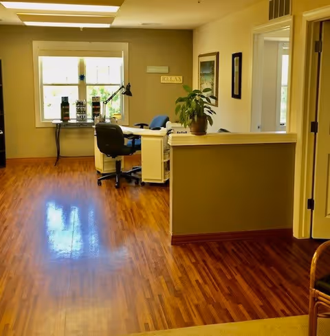 Interior view of a well-lit office or reception area with wooden flooring, a desk with a black office chair, a potted plant on a half wall, and a window letting in natural light. The walls are decorated with framed pictures and a sign that reads 'RELAX'.