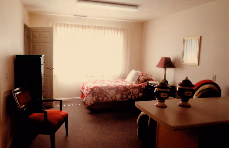 A bedroom with a bed covered in a red floral bedspread positioned near a window with sheer curtains. There is a wooden chair with a red cushion on the left side, a tall dresser near the door, a nightstand with a lamp beside the bed, and a table with decorative items in the foreground.