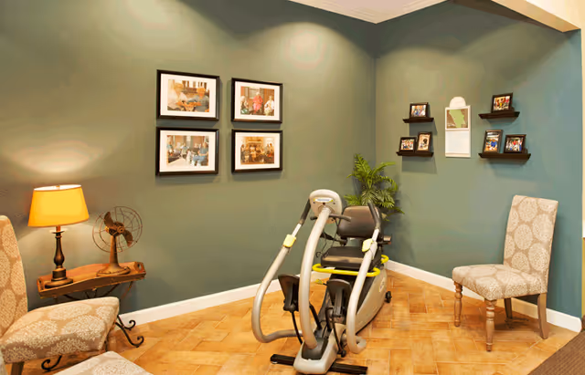 A cozy room with green walls featuring a recumbent exercise bike in the center. There are two patterned chairs on either side of the room, a small wooden table with a lamp and a vintage fan, framed photos on the wall, and small shelves holding picture frames and a calendar. A potted plant is placed in the corner.