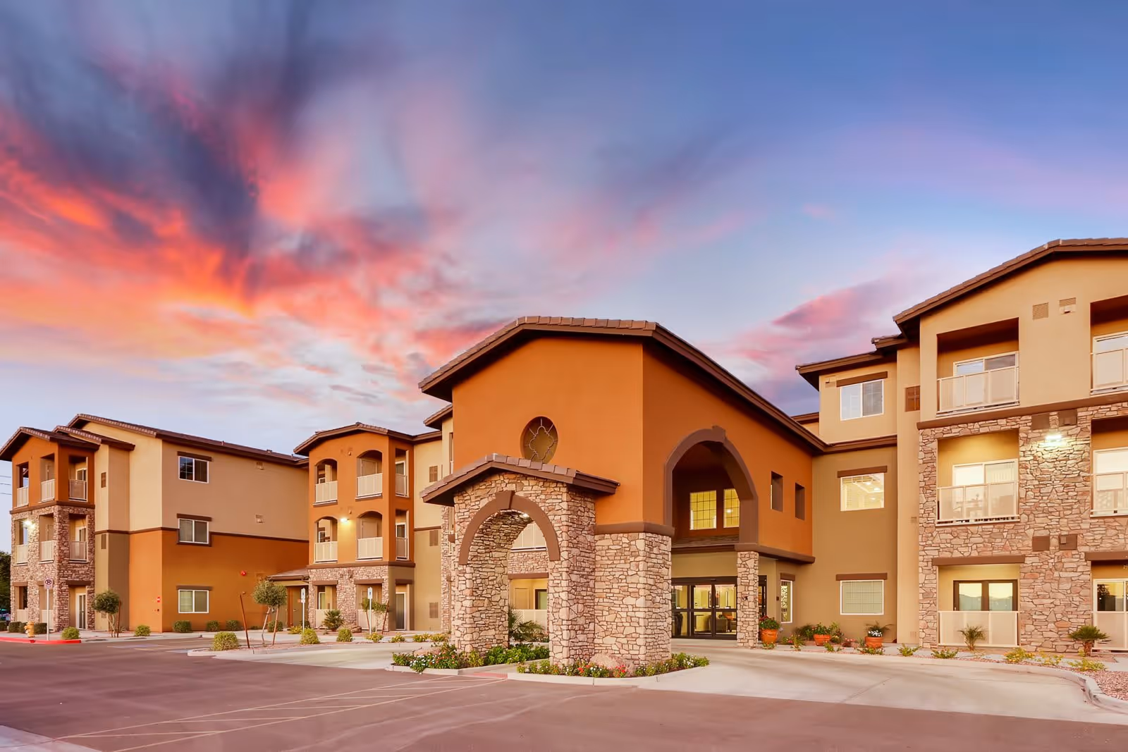 Exterior view of a multi-story senior living facility building with stone and stucco facade under a colorful sunset sky with pink and purple clouds.