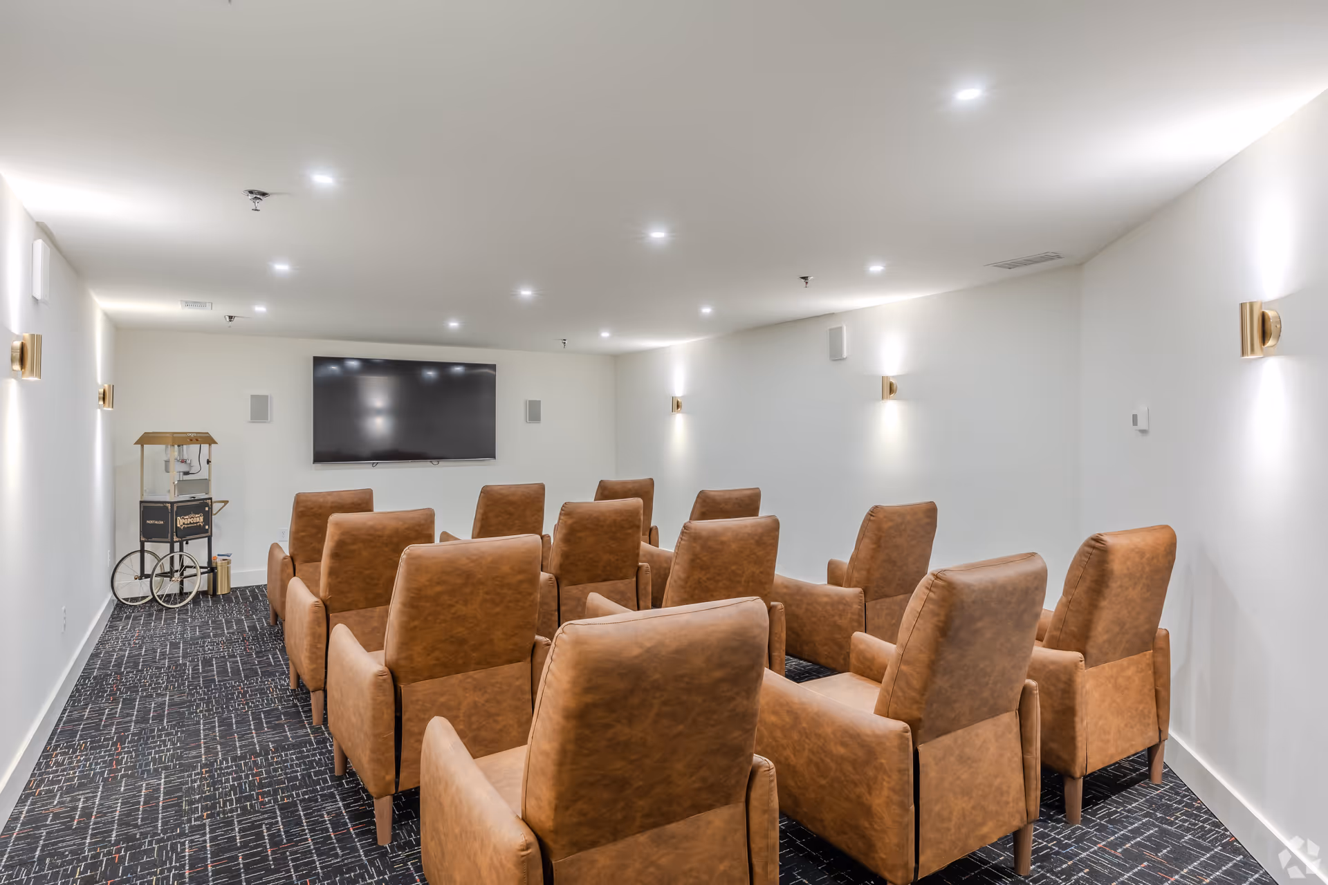 A small theater room with two rows of brown leather armchairs facing a large flat-screen TV mounted on a white wall. The room has a dark patterned carpet, white walls with modern wall sconces, and a popcorn machine on a cart in the corner.