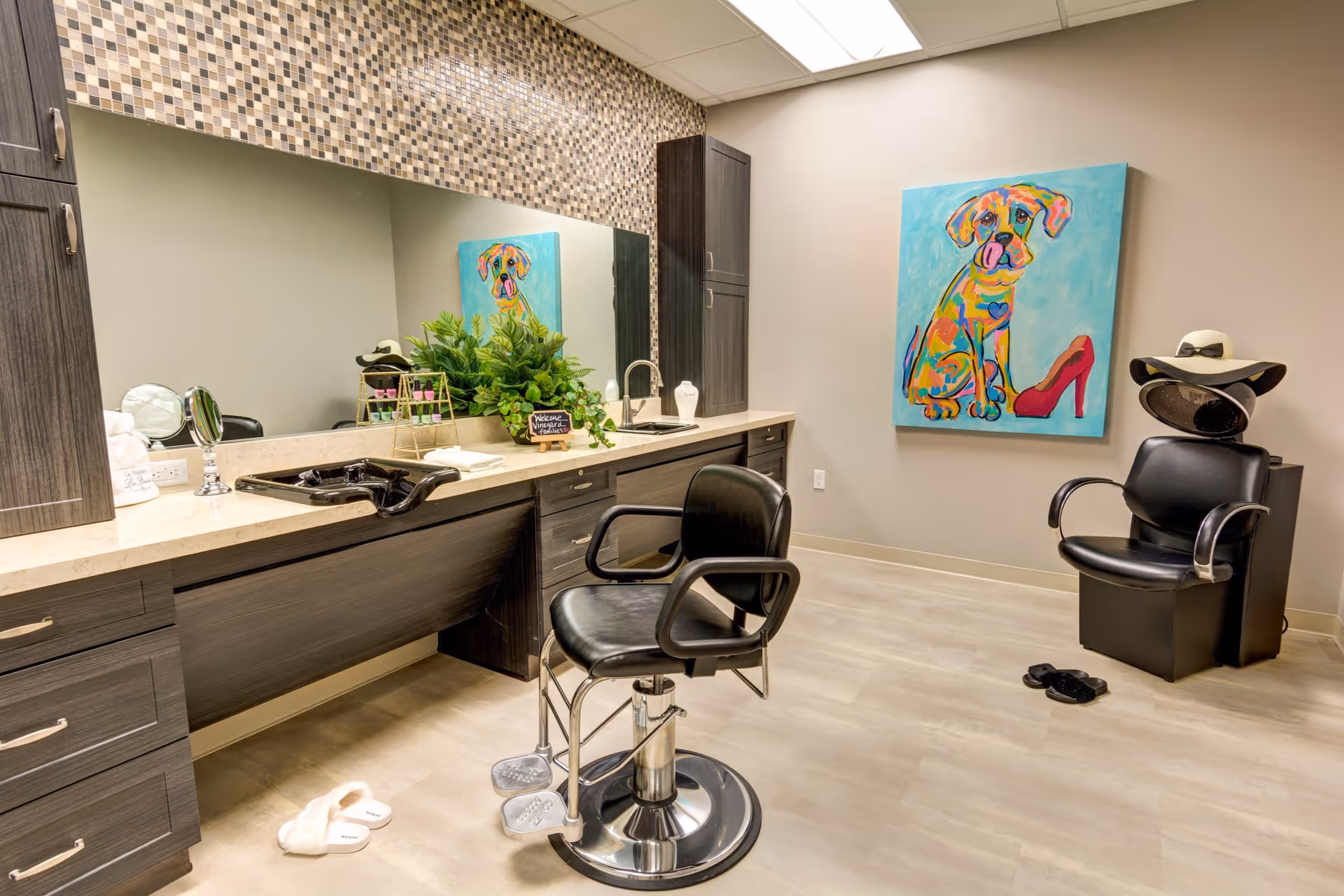 A modern salon room with a black salon chair in front of a long counter with a sink and large mirror. The counter has dark wood cabinets and a mosaic tile backsplash. On the wall is a colorful painting of a dog next to a red high heel shoe. There is another black chair with a hair dryer hood and a hat on top. Slippers are on the floor near the chairs.
