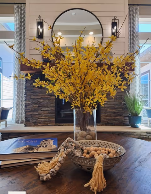 A living room table vignette with a tall glass vase of yellow branches and a bowl of decorative nuts and tassels in front of a stone fireplace and round mirror.