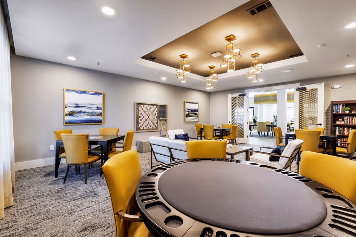 Communal lounge with yellow chairs, a round card table in the foreground, sofas and pendant lights in a bright seating area.